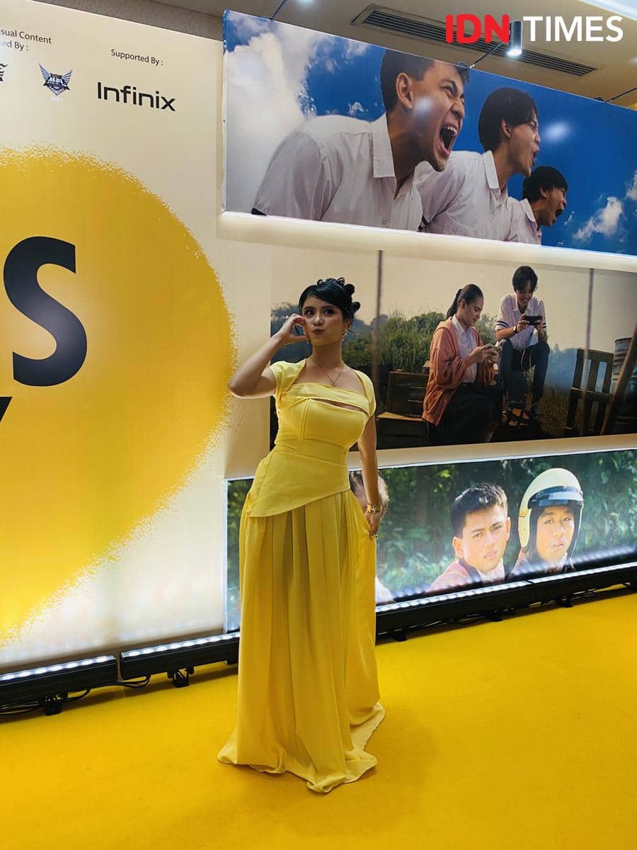 Seorang perempuan mengenakan gaun kuning elegan berpose di karpet kuning acara pemutaran film Nobody Loves Kay dengan latar poster film.