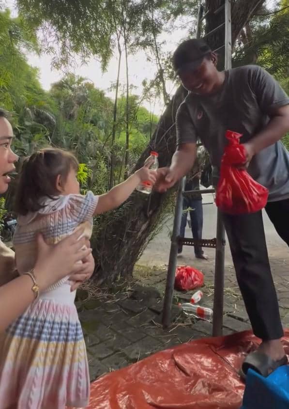 Seorang anak kecil menyerahkan botol minuman kepada pria yang memegang kantong plastik merah di area luar dengan pepohonan hijau di sekitarnya.