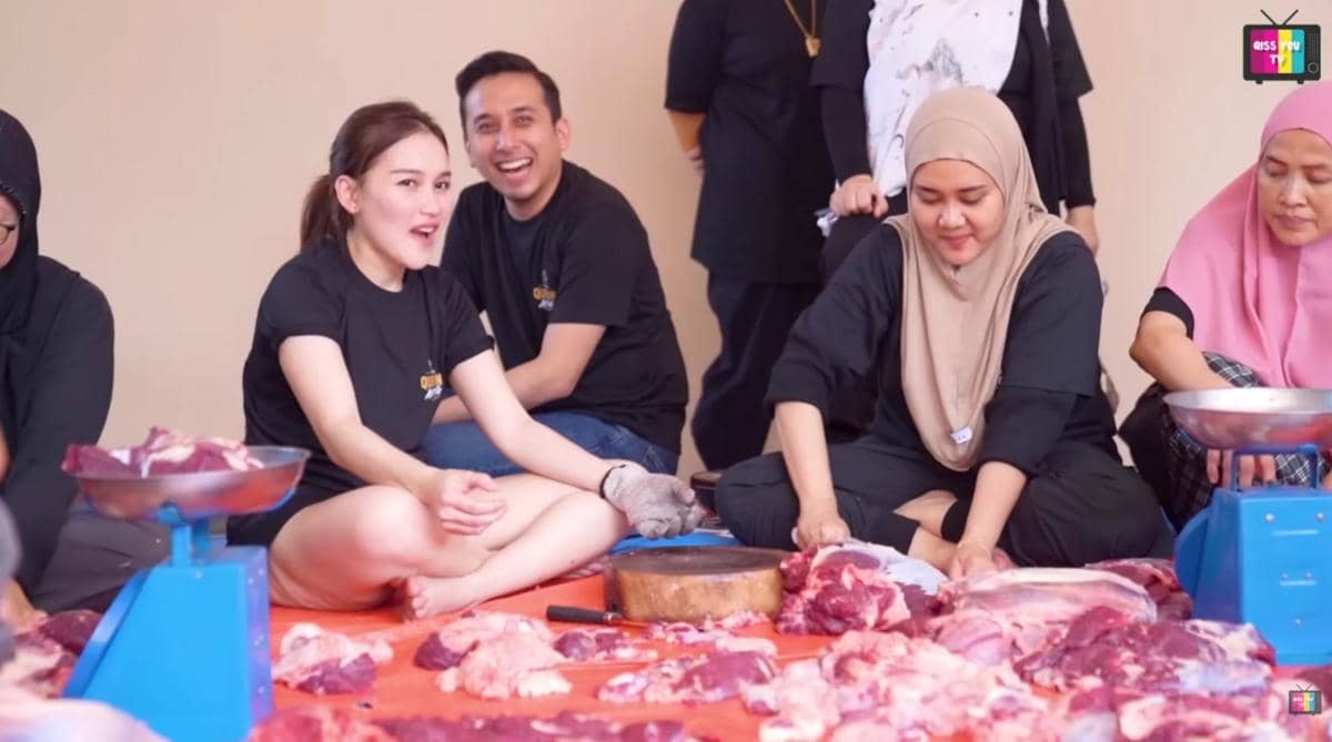 Beberapa orang mengenakan pakaian hitam sedang memotong dan menimbang daging sapi kurban di atas alas oranye dengan suasana santai.