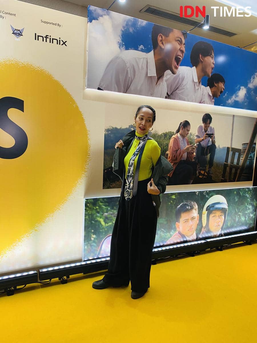 Seorang tamu berpose di karpet kuning acara pemutaran film Nobody Loves Kay dengan latar poster film dan logo sponsor.
