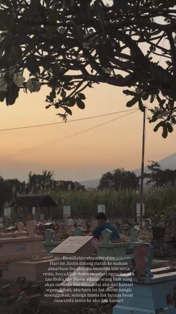 Seorang pria duduk di samping makam di pemakaman saat matahari terbenam, dengan pepohonan dan latar pegunungan di kejauhan.