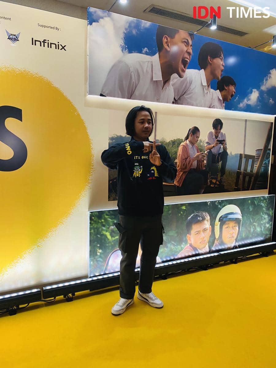 Seseorang berpose di karpet kuning acara pemutaran film Nobody Loves Kay dengan latar poster film dan logo sponsor Infinix.