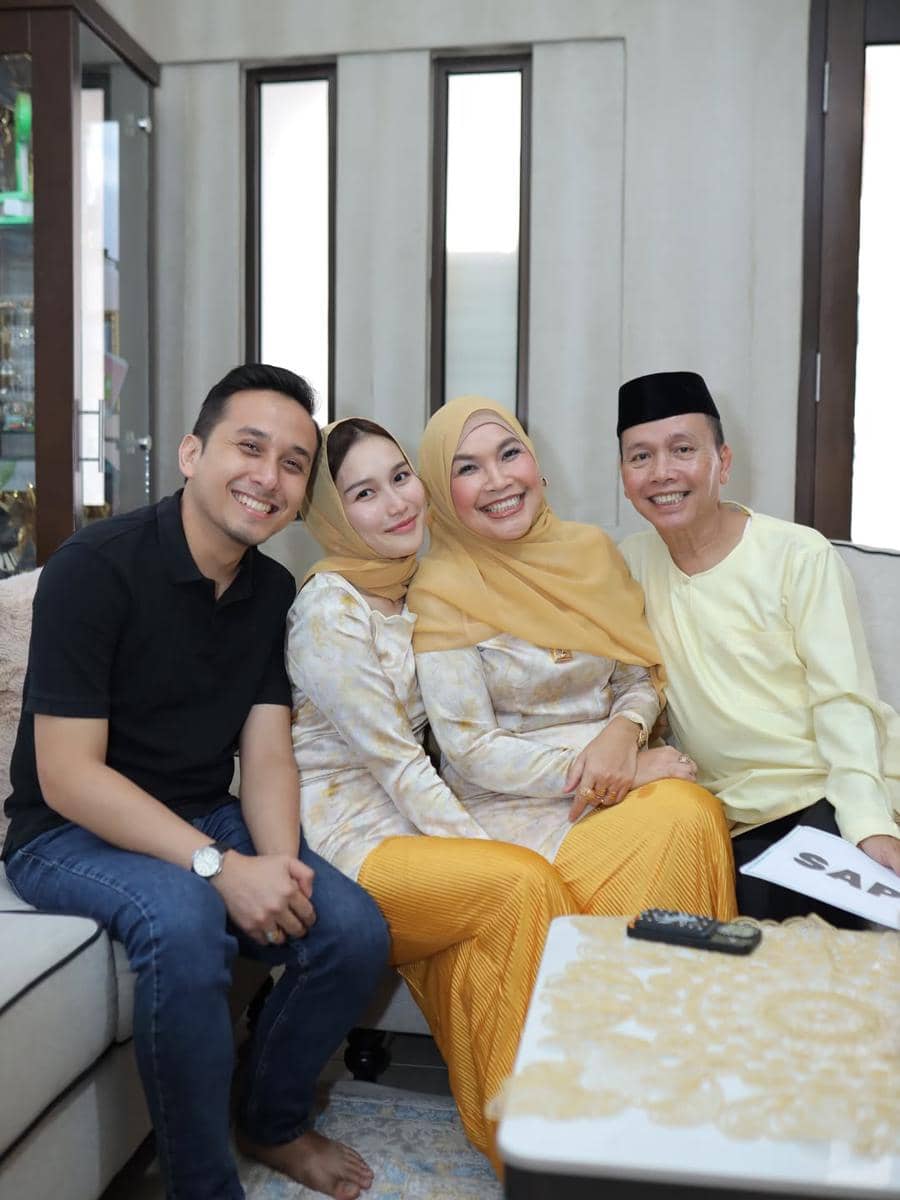 Empat orang duduk bersama di ruang tamu, dua wanita mengenakan hijab kuning dan dua pria tersenyum ke arah kamera dengan suasana hangat.