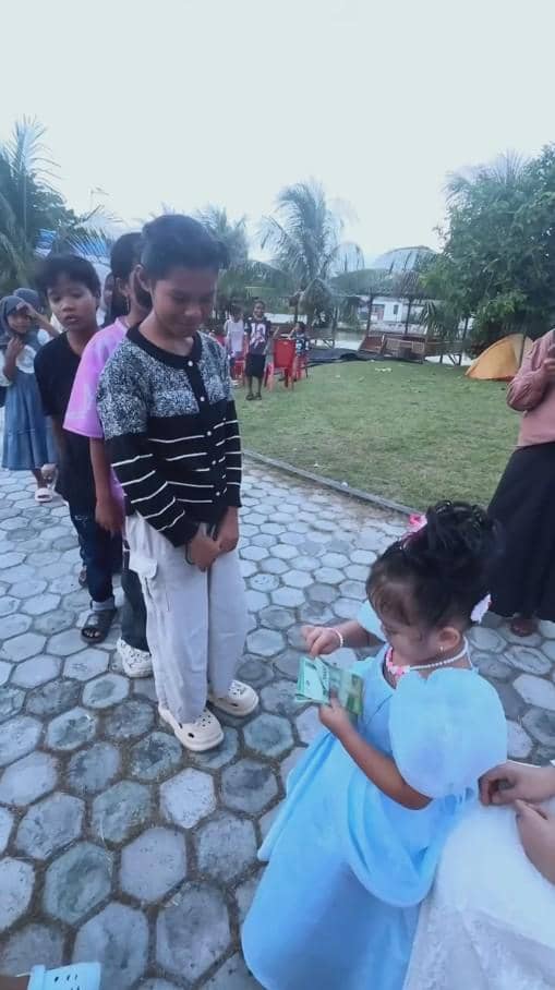 Seorang anak kecil mengenakan gaun biru membagikan uang kepada anak-anak lain yang berbaris di area terbuka dengan latar pepohonan.