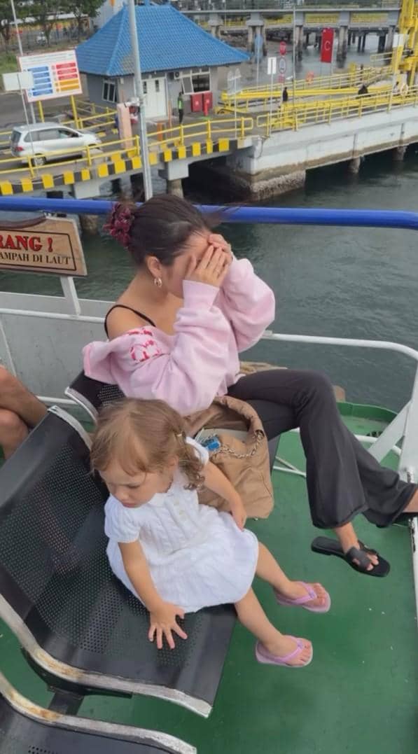 Seorang perempuan mengenakan sweater merah muda duduk di kapal bersama seorang anak kecil bergaun putih di dekat dermaga.
