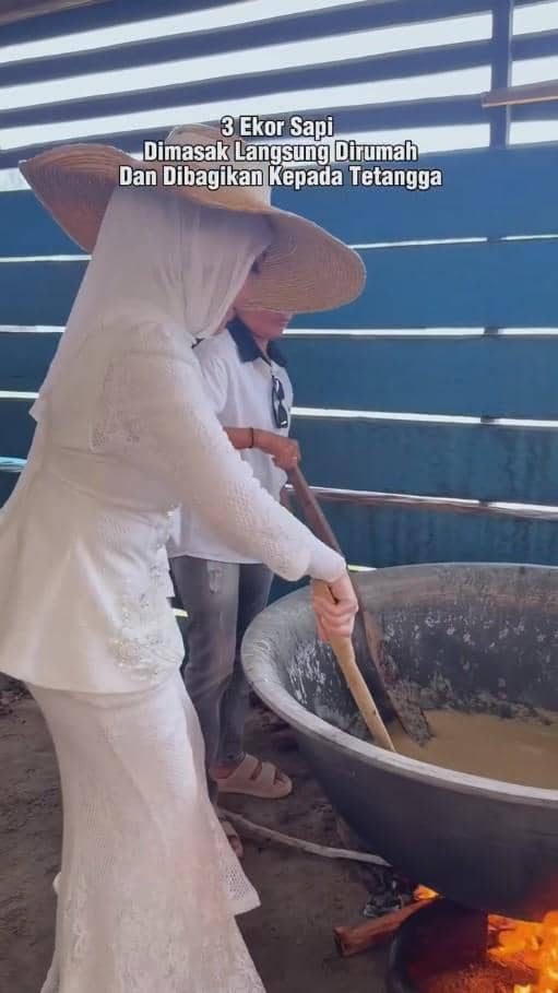 Seorang wanita berhijab mengenakan kebaya putih dan topi lebar sedang mengaduk masakan besar di kuali di dapur terbuka.