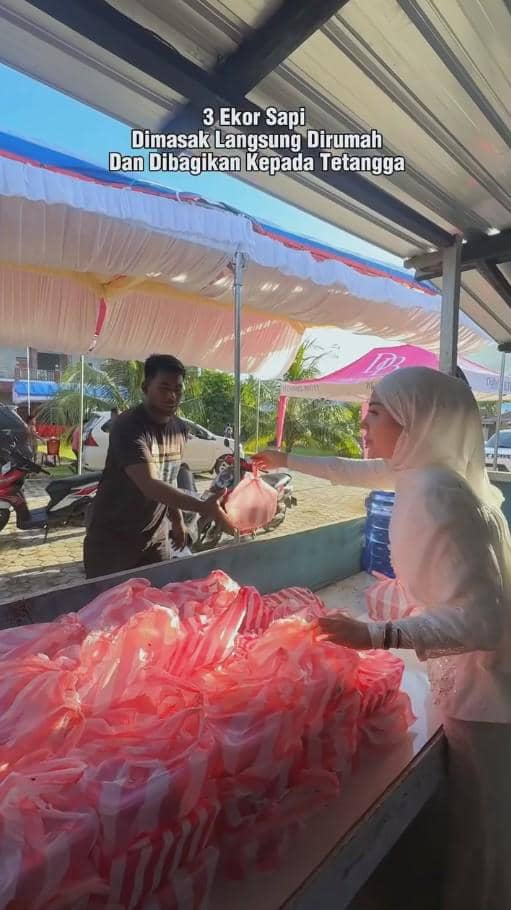 Seorang wanita berhijab membagikan daging kurban dalam kantong plastik merah muda kepada seorang pria di area terbuka.