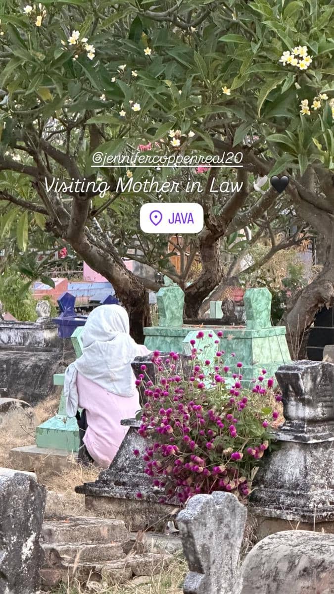 Seorang perempuan berhijab duduk di area pemakaman dengan nisan berwarna hijau dan bunga ungu di sekitarnya di Pulau Jawa.