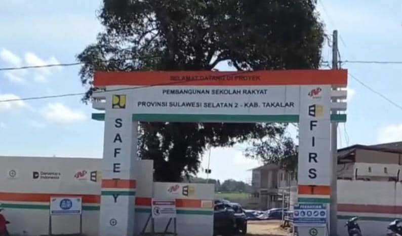 Gerbang proyek pembangunan sekolah rakyat di Kabupaten Takalar, Sulawesi Selatan, dengan tulisan keselamatan kerja di area pintu masuk.