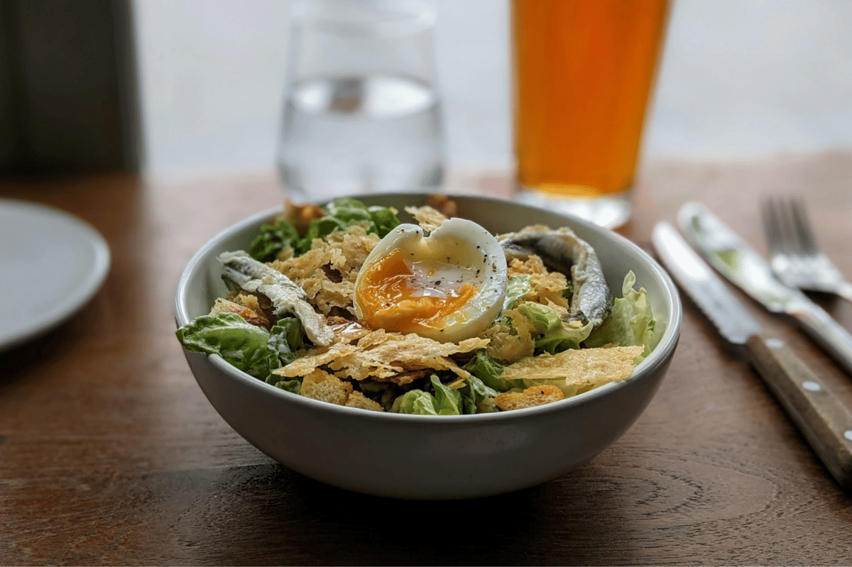Semangkuk salad telur gaya Prancis dengan telur setengah matang, sayuran hijau segar, ikan teri, dan taburan keripik di atas meja kayu.