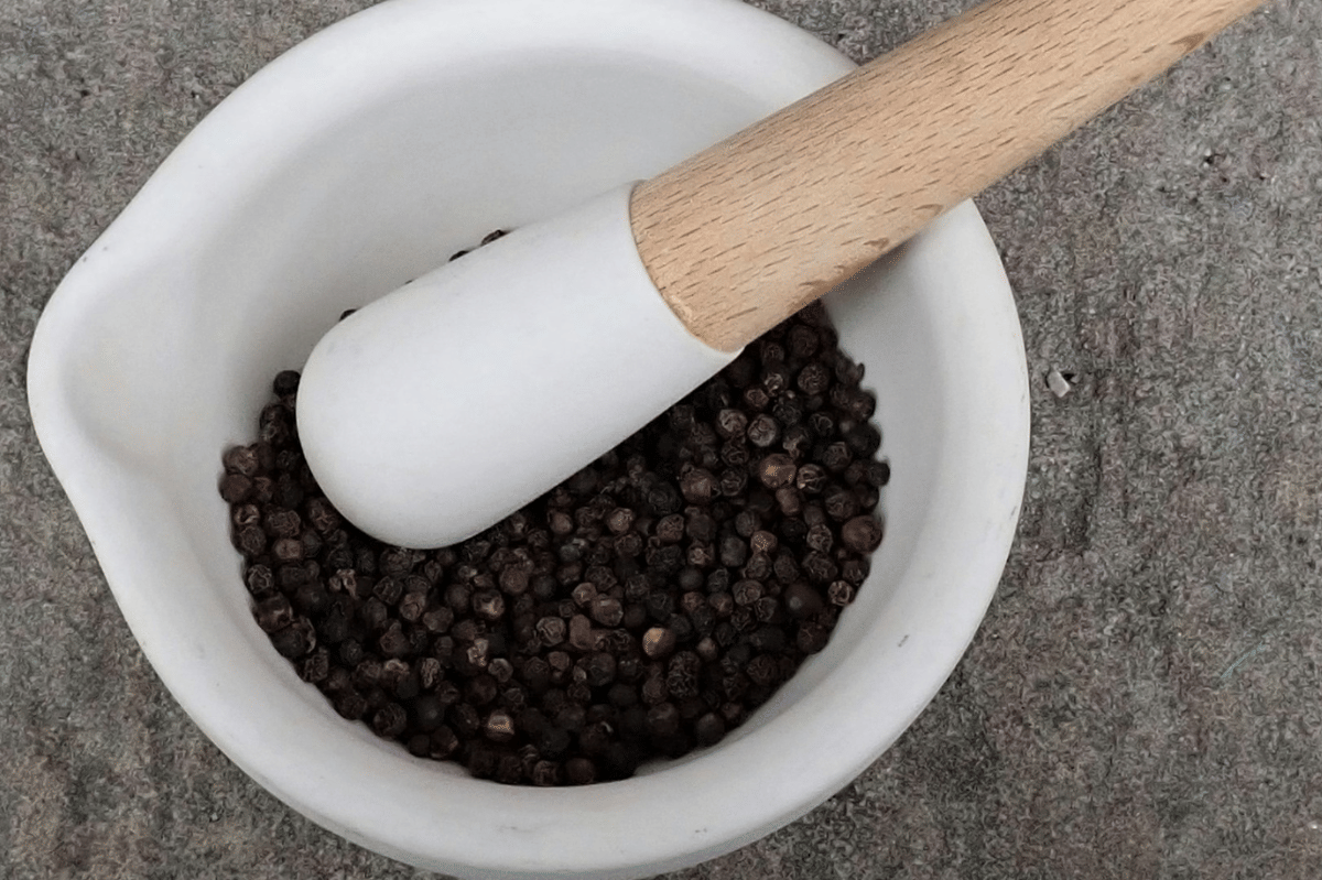 Butiran lada hitam kering di dalam cobek putih dengan ulekan kayu di atas permukaan batu abu-abu.