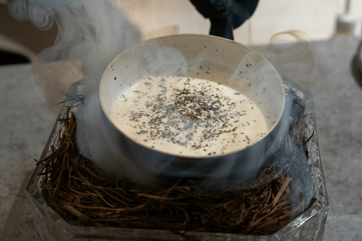 Semangkuk saus peppercorn berwarna krem dengan taburan lada hitam di atasnya, diletakkan di atas wadah berisi ranting dan asap tipis.