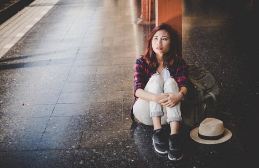 Seorang perempuan duduk bersandar di lantai stasiun dengan ekspresi termenung, di sampingnya terdapat ransel dan topi jerami.