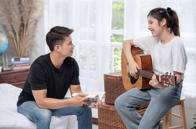 Pasangan muda duduk di kamar bercahaya alami, berbincang sambil tersenyum, salah satunya memegang gitar akustik.
