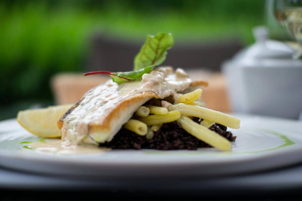 Hidangan ikan panggang dengan saus peppercorn disajikan di atas kacang panjang dan nasi hitam dengan hiasan daun segar.