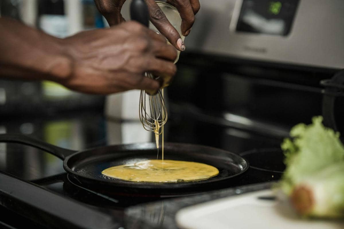 Seseorang menuangkan adonan telur kocok ke dalam wajan panas di dapur untuk membuat omelet dengan alat pengocok di tangan.