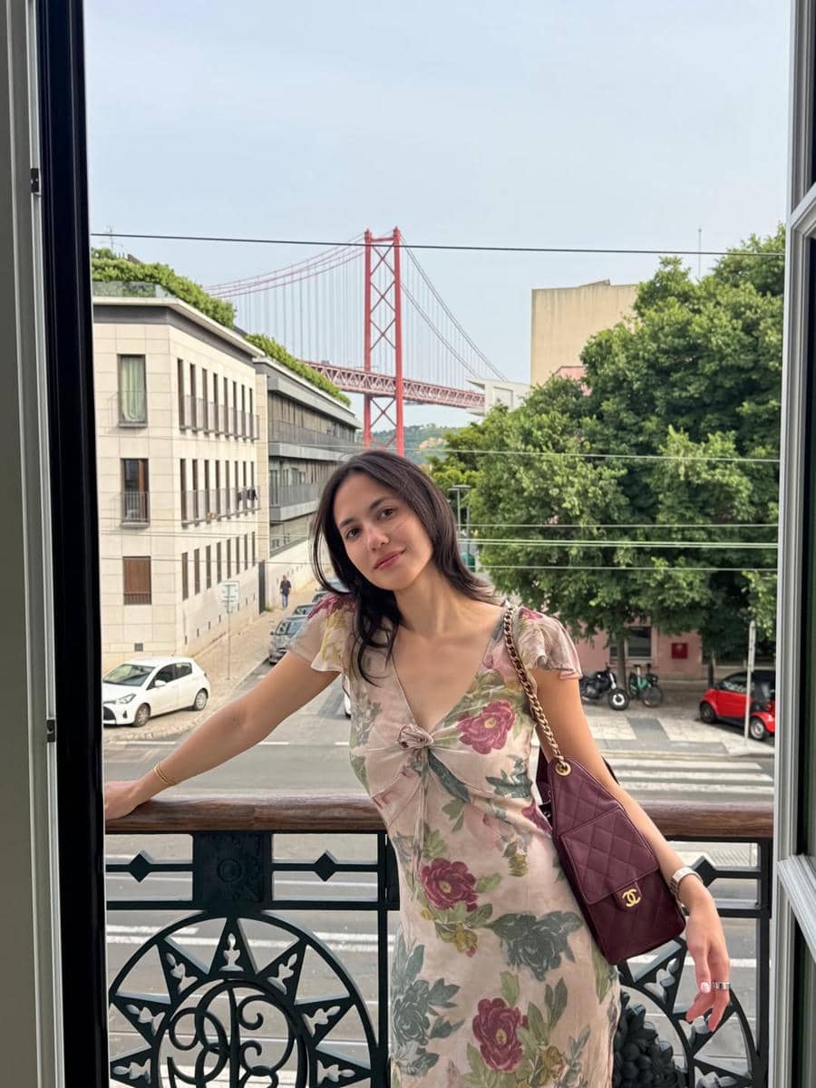 Seorang perempuan mengenakan gaun bermotif bunga berdiri di balkon dengan latar jembatan merah dan bangunan kota Lisbon.