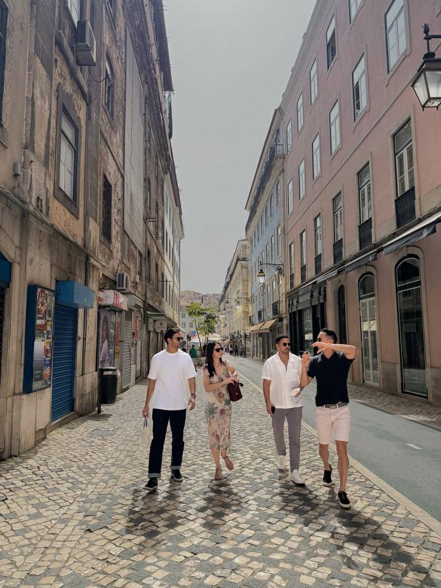 Empat orang berjalan santai di jalan berbatu dengan bangunan bergaya klasik di Kota Lisbon pada siang hari yang cerah.
