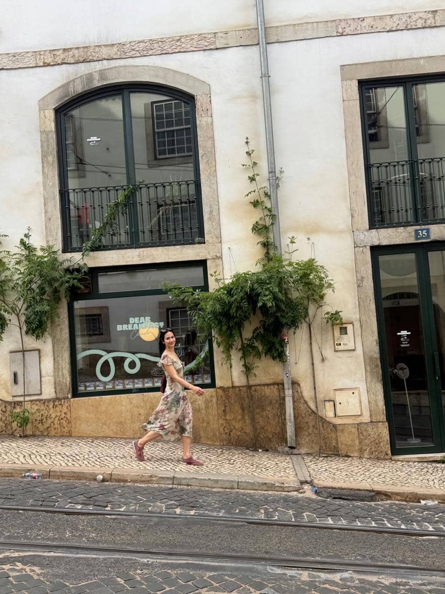 Seorang wanita mengenakan gaun bermotif bunga berjalan di trotoar berbatu di depan bangunan bergaya Eropa di Kota Lisbon.