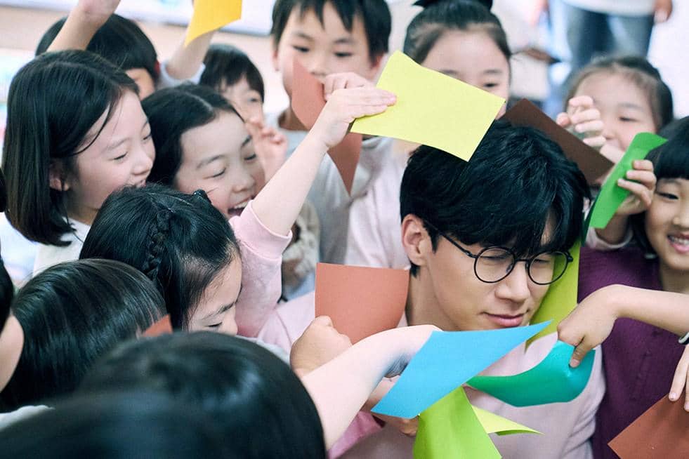 Seorang pria berkacamata dikelilingi anak-anak yang tersenyum sambil memegang kertas warna-warni di ruang kelas.