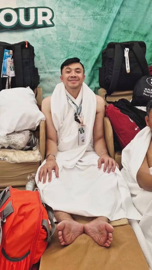 Seorang pria mengenakan kain ihram duduk santai di kursi dengan latar belakang tas dan perlengkapan haji di sekitarnya.
