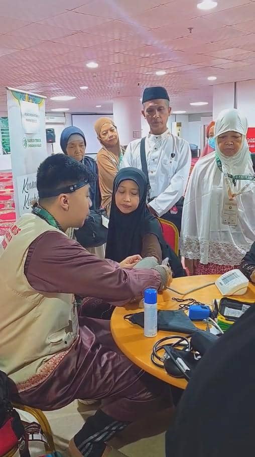 Seorang petugas medis memeriksa tekanan darah jamaah haji perempuan di ruang pelayanan kesehatan dengan beberapa orang menunggu di belakang.