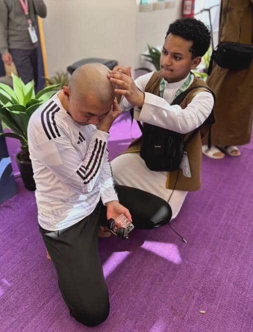 Seorang pria mencukur rambut seorang jamaah yang sedang berlutut di atas karpet ungu dalam suasana ibadah haji.