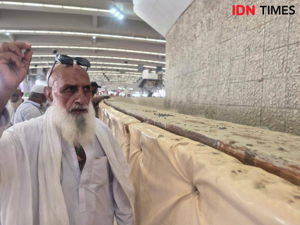Seorang jemaah haji berpakaian ihram putih sedang melontar jumrah Aqobah di area Jamarat, dengan latar dinding batu besar.