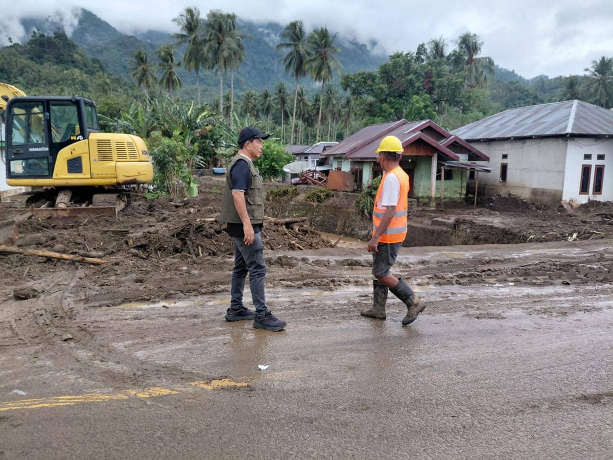 Dua petugas berdiri di area berlumpur pascabanjir di Kecamatan Bolaang, Bolmong, dengan alat berat dan rumah warga di latar belakang.
