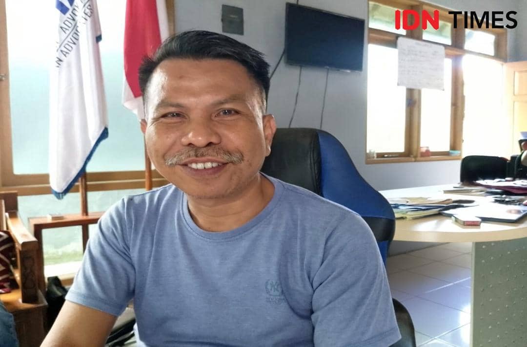 Seorang pria mengenakan kaus biru tersenyum di dalam ruangan kantor dengan meja kerja, bendera, dan jendela di latar belakang.