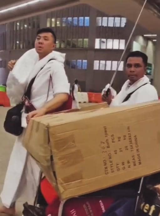 Dua pria berpakaian ihram membawa koper besar dan kardus di area bandara dengan suasana malam yang ramai dan terang.