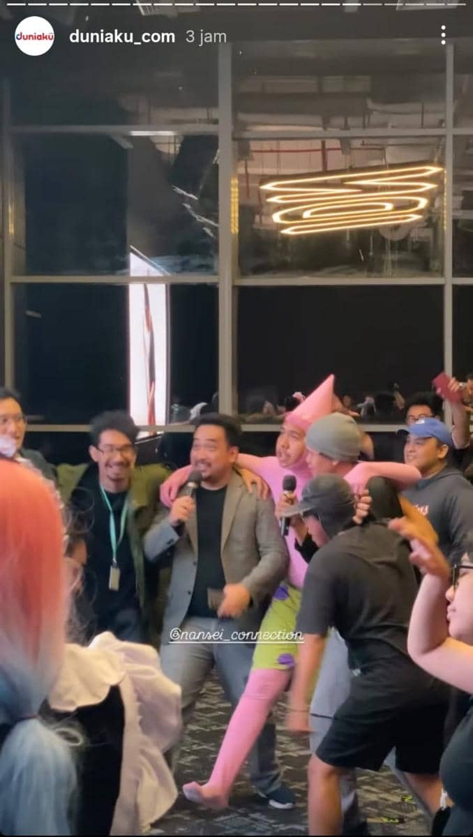 Beberapa orang bernyanyi bersama dengan mikrofon di acara karaoke Warga di The Plaza, IDN HQ, dengan suasana meriah dan kostum unik.