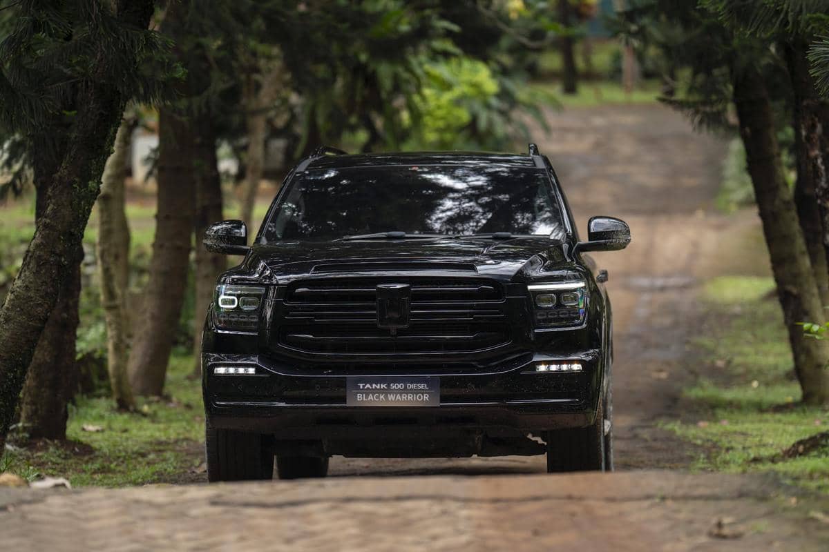 SUV hitam GWM Tank 500 Diesel Black Warrior melaju di jalan taman dengan pepohonan rindang di sekitarnya.