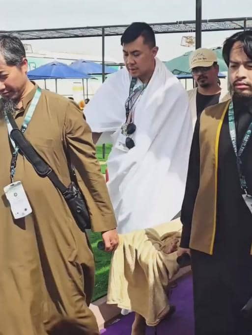 Beberapa pria berpakaian ihram dan gamis berjalan bersama di area terbuka dengan latar tenda dan payung saat kegiatan haji.