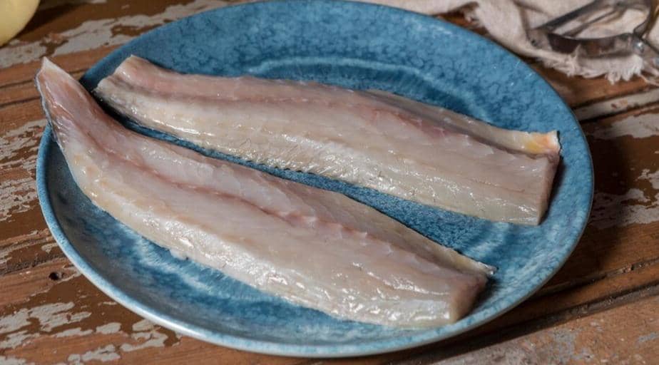 Dua potong fillet ikan tenggiri mentah diletakkan di atas piring biru di atas meja kayu dengan pencahayaan alami.