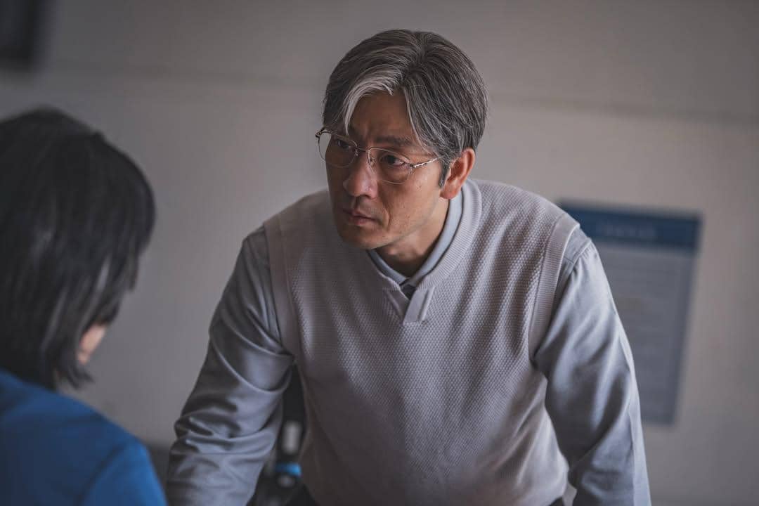 Seorang pria berambut abu-abu mengenakan kacamata dan sweter abu-abu sedang berbicara serius dengan seseorang di depannya dalam adegan drama The Scarecrow.