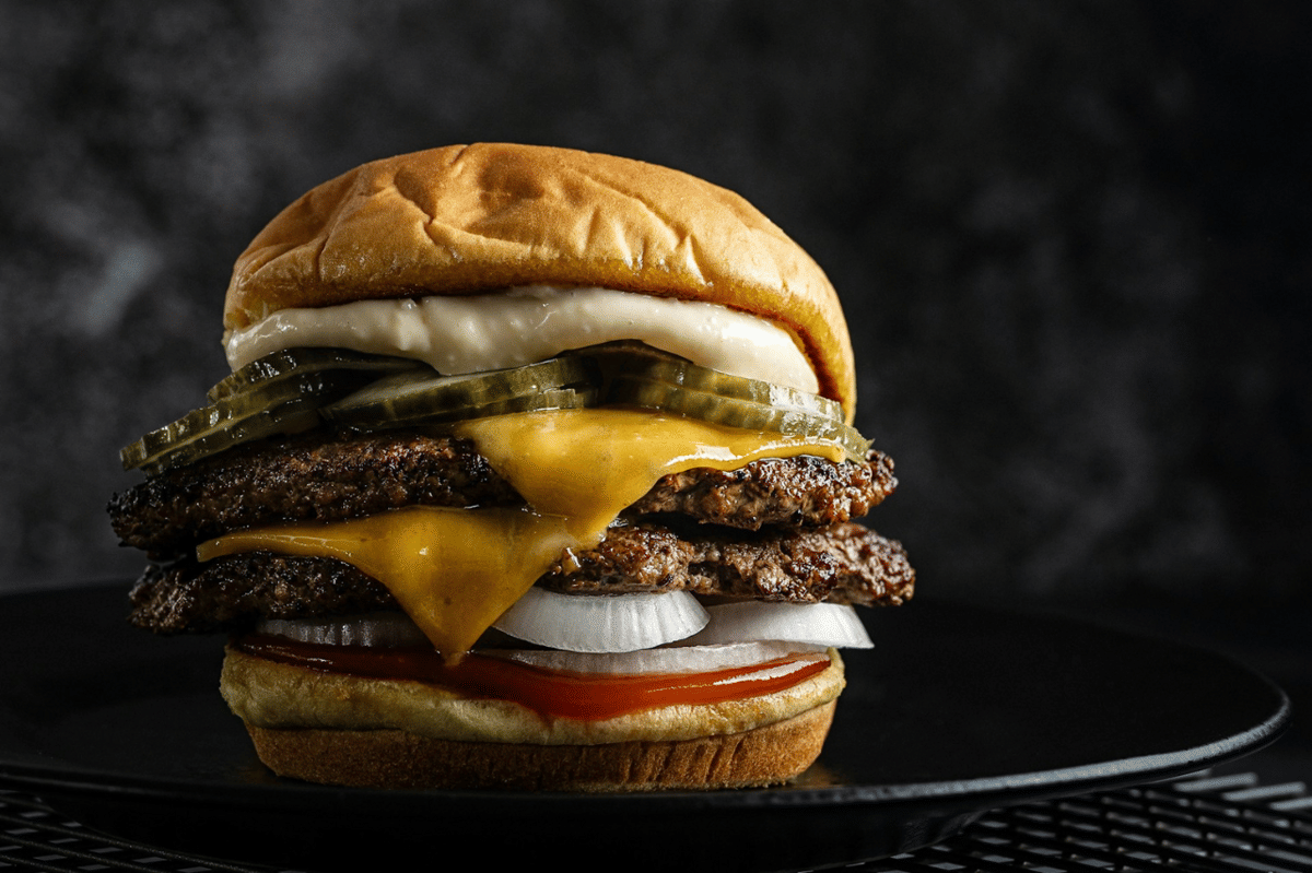 Double cheeseburger dengan dua lapis daging sapi, keju leleh, acar, bawang, saus tomat, dan roti lembut di atas piring hitam.