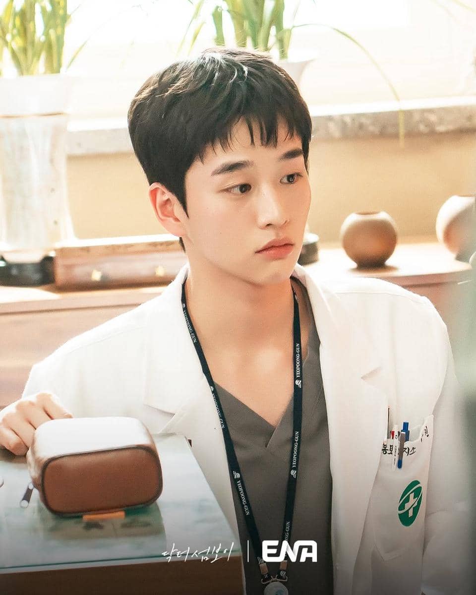Seorang aktor mengenakan jas dokter putih dengan latar ruangan terang dan tanaman hias di belakangnya dalam adegan serial Doctor on the Edge.