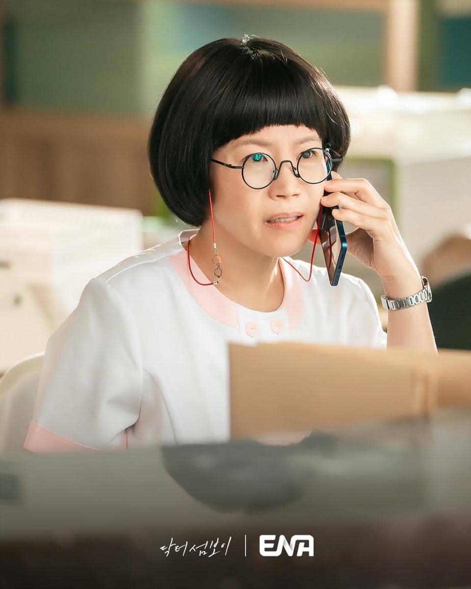 Seorang wanita berambut bob mengenakan kacamata dan seragam medis putih muda sedang berbicara di telepon di sebuah ruangan kerja.