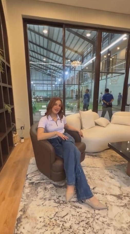 Seorang wanita duduk santai di kursi ruang lounge dengan latar belakang lapangan padel indoor yang terlihat melalui dinding kaca besar.