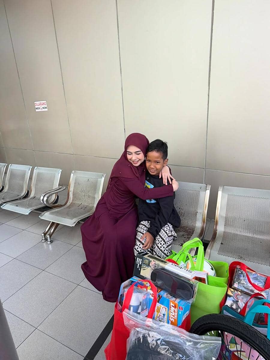 Seorang wanita berhijab ungu memeluk seorang anak laki-laki di kursi ruang tunggu dengan beberapa tas belanja berisi mainan di depannya.