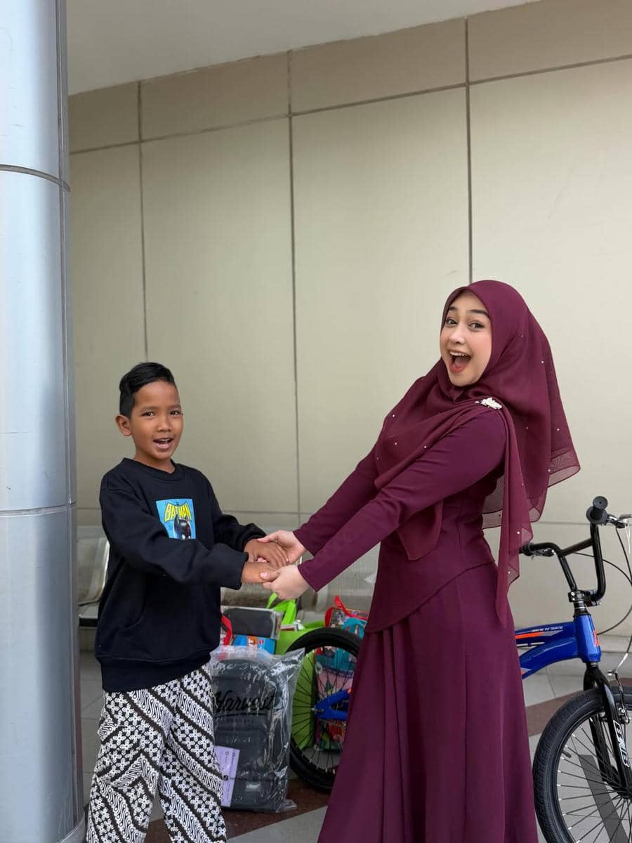 Seorang wanita berhijab ungu tersenyum sambil memegang tangan seorang anak laki-laki di depan dinding luar gedung dengan beberapa barang belanjaan dan sepeda di belakang mereka.