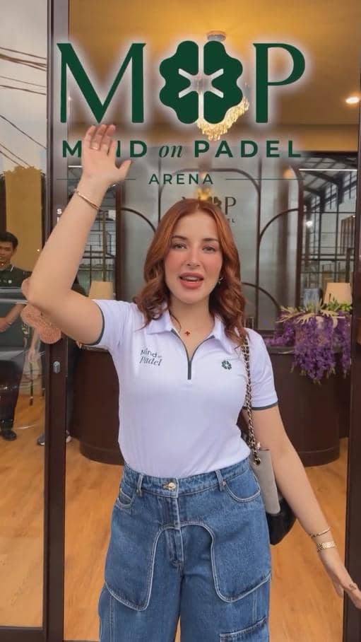 Seorang perempuan mengenakan kaus putih dan celana jeans berdiri di depan pintu masuk Mind on Padel Arena sambil melambaikan tangan.