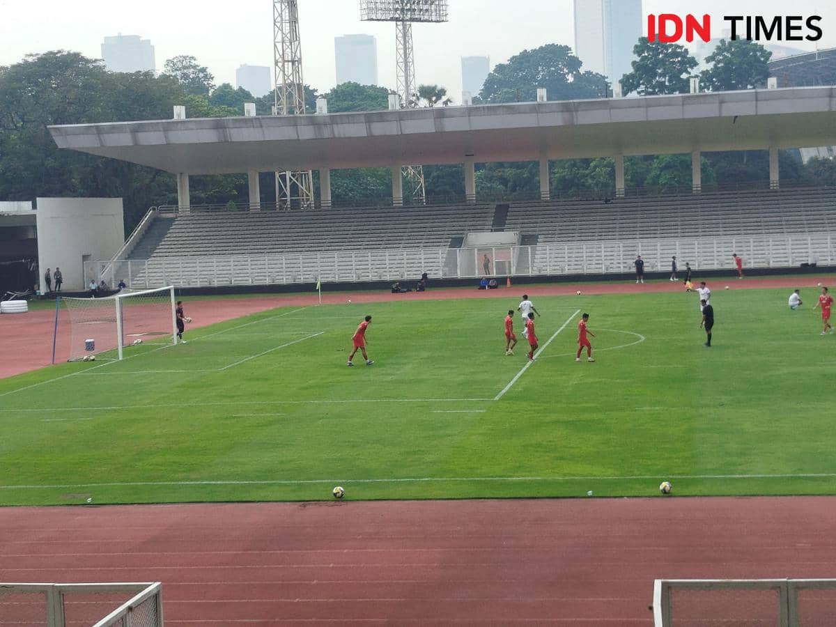 Pemain Timnas Indonesia berlatih di Stadion Madya dengan latar tribun kosong dan gedung-gedung tinggi di kejauhan.