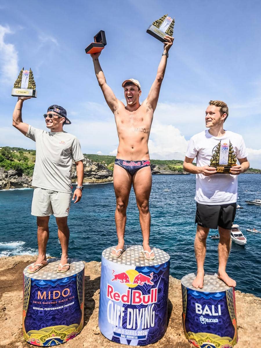 Tiga atlet berdiri di podium tepi tebing sambil mengangkat trofi pada ajang Red Bull Cliff Diving 2026 di Bali dengan latar laut biru.