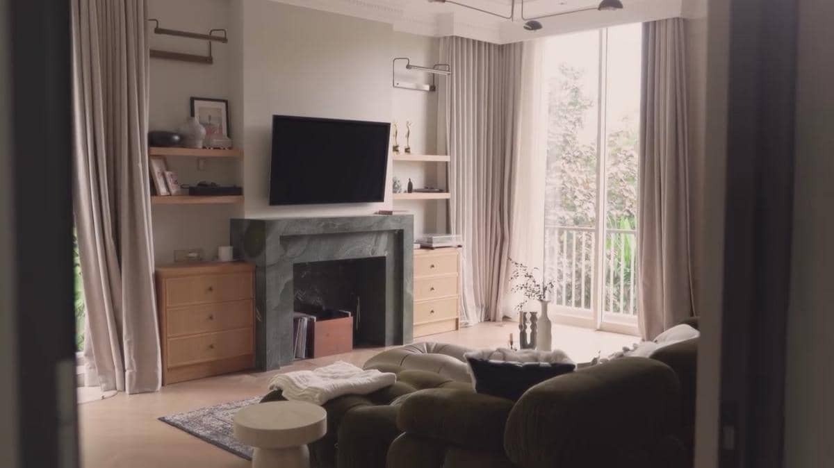 Ruang tamu rumah mewah dengan sofa hijau, perapian marmer, televisi di dinding, dan jendela besar dengan tirai panjang.