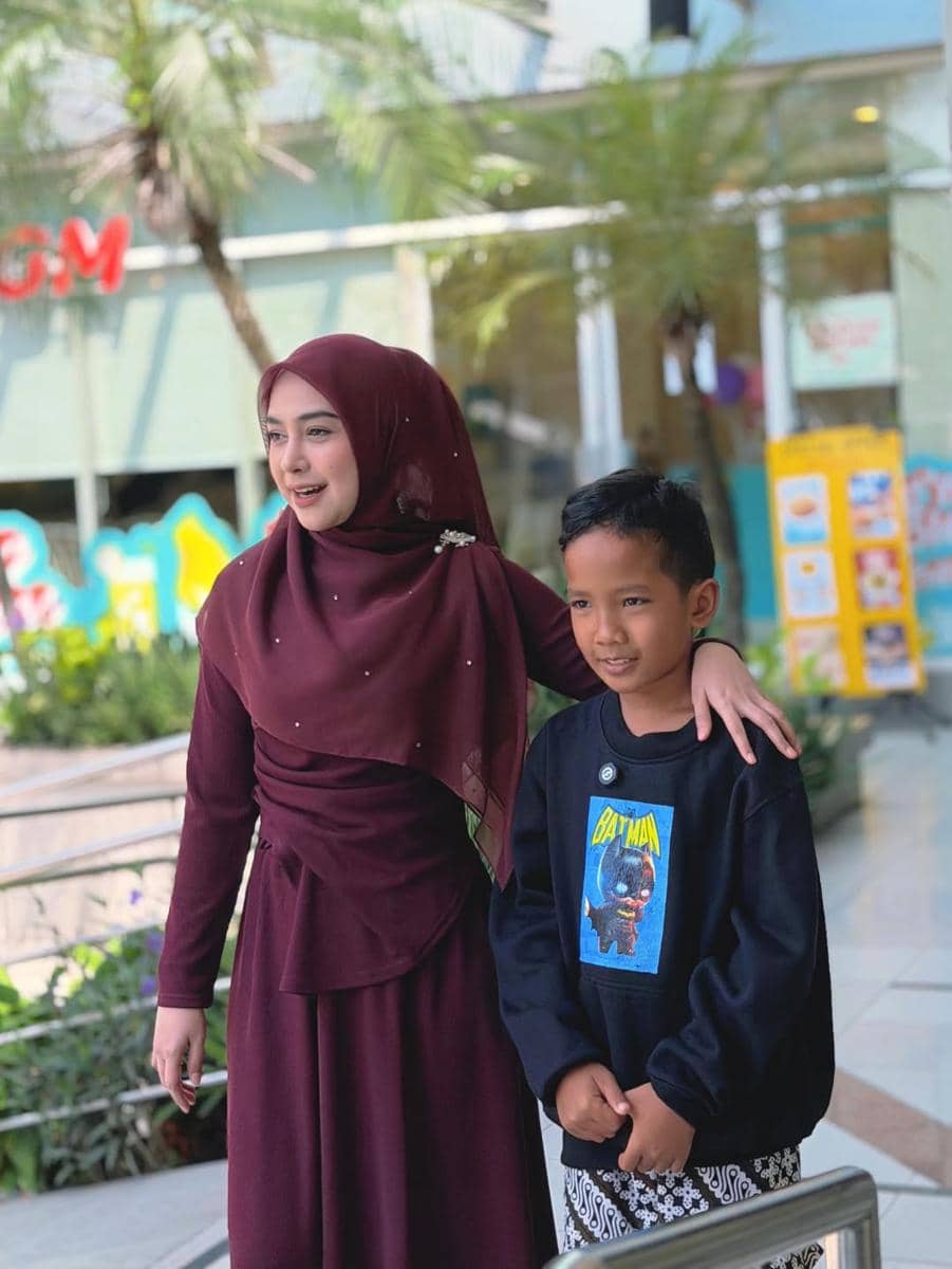 Seorang wanita berhijab berwarna marun tersenyum sambil merangkul seorang anak laki-laki kecil yang mengenakan sweater hitam bergambar Batman di area luar pusat perbelanjaan.