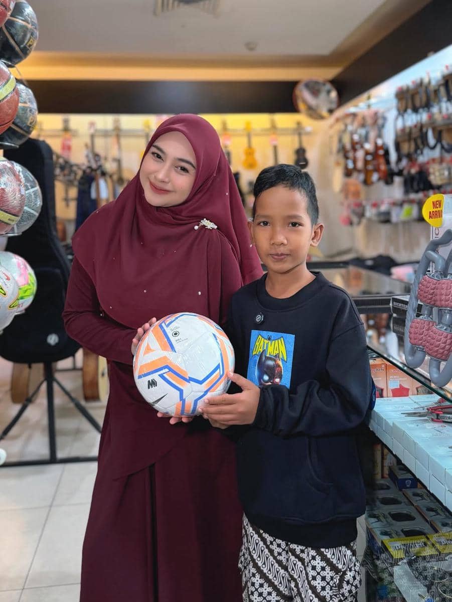 Seorang wanita berhijab warna marun tersenyum sambil berdiri di samping anak laki-laki yang memegang bola di dalam toko alat musik.