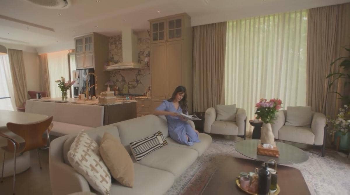 Seorang wanita duduk di sofa ruang tamu rumah mewah dengan desain modern, pencahayaan alami, dan dekorasi elegan bernuansa hangat.