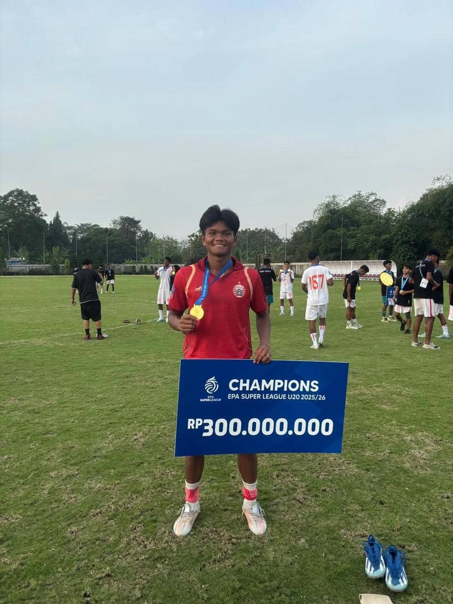 Seorang pemain sepak bola muda mengenakan seragam merah memegang papan hadiah juara EPA Super League U20 senilai Rp300 juta di lapangan.
